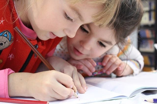 a young girl child writing in a book with a pencil with concentration while another looks on  